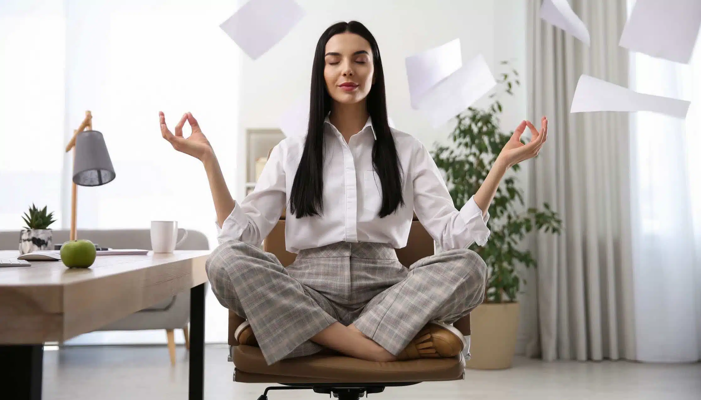 Frau sitzt im Büro im Schneidersitz auf dem Stuhl und meditiert mit geschlossenen Augen sowie entspannten Händen. Blätter fliegen im Hintergrund. Fokus auf Ruhe und Stressabbau.