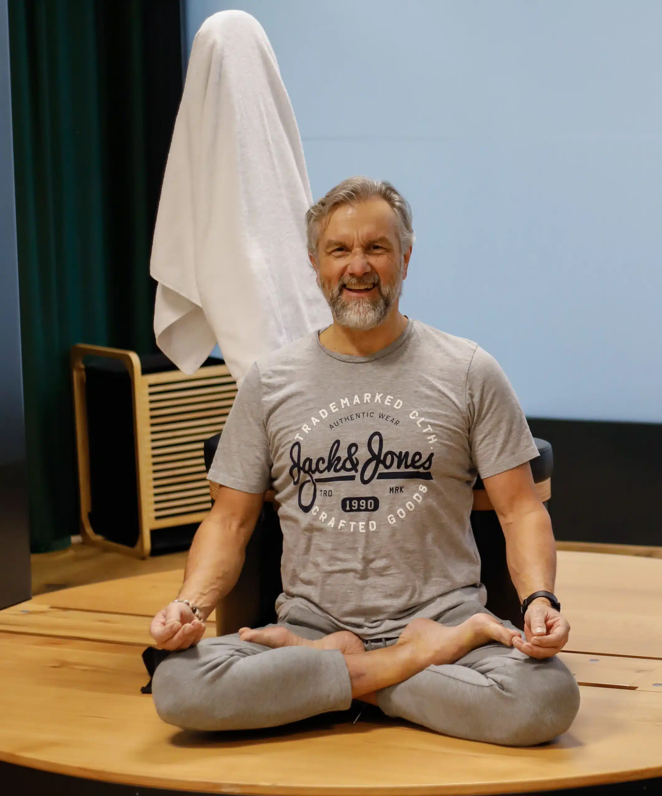 Thomas Gloor sitzt im Lotussitz und lächelt. Ruhige Haltung in einem Innenraum.