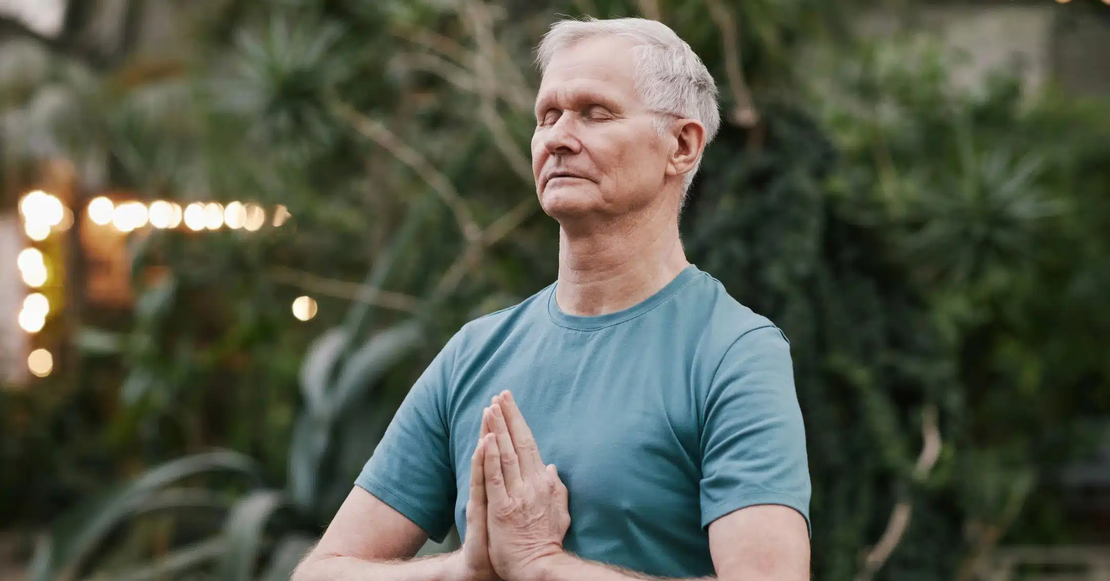 Executive Coaching Zug. Älterer Mann sitzt ruhig im Freien, mit geschlossenen Augen und gefalteten Händen vor der Brust.